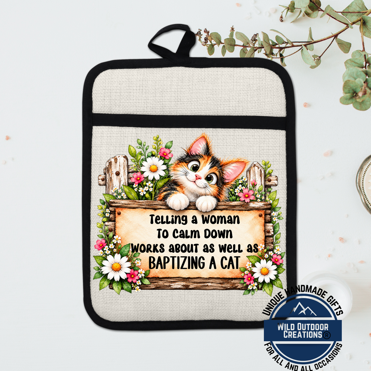 A beige oven mitt with a cat illustration and text, placed on a white surface next to a plant.