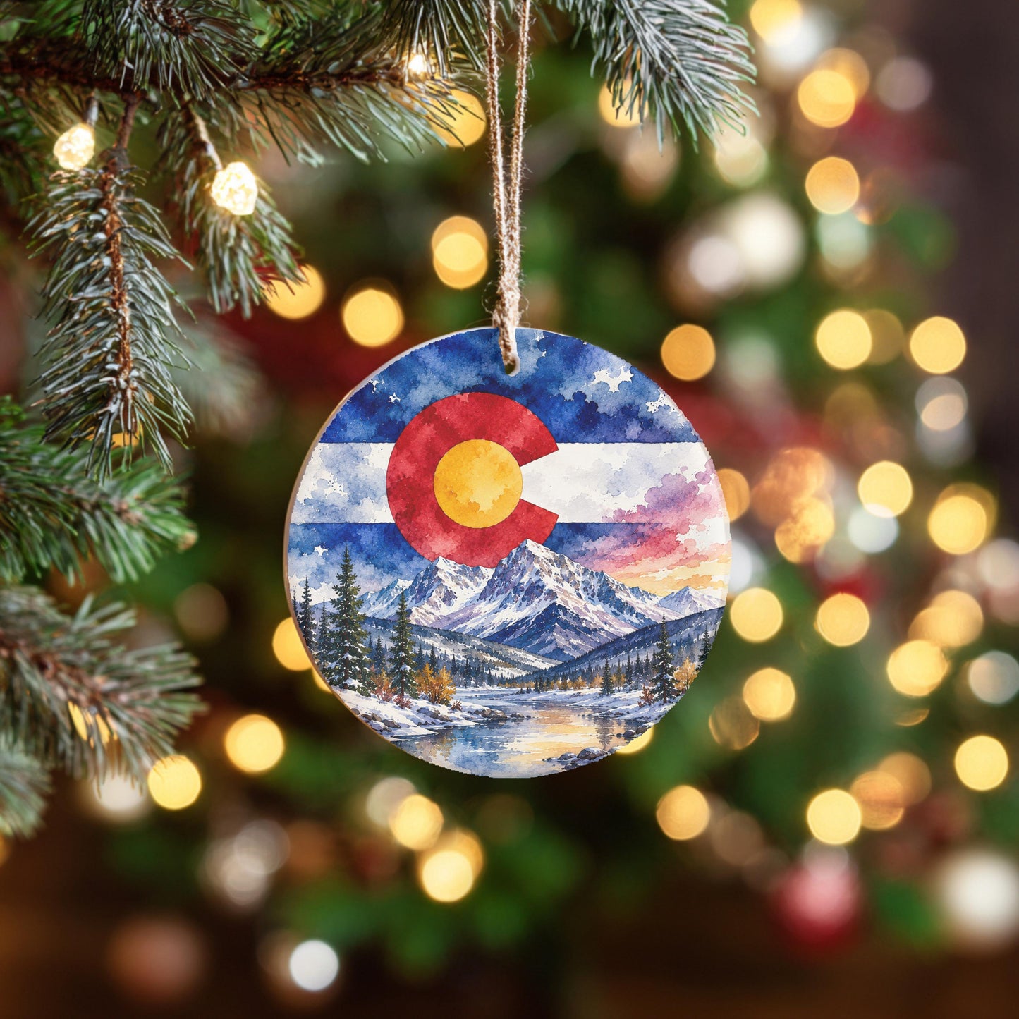 A round ornament hanging from a Christmas tree, featuring a painting of a snowy mountain landscape with a Colorado flag in the center.