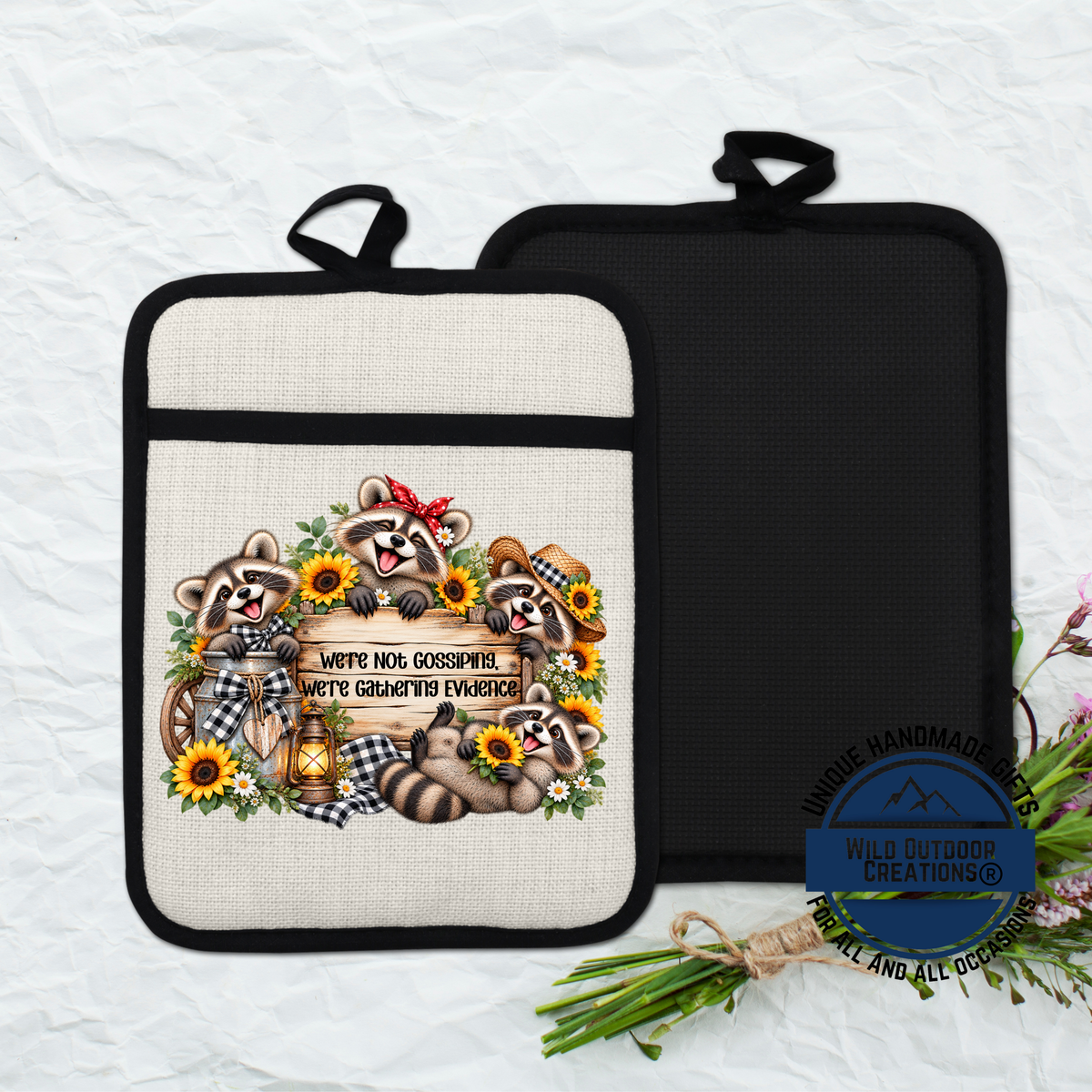 a beige pot holder with a black handle, decorated with a whimsical illustration of raccoons surrounded by flowers and sunflowers.