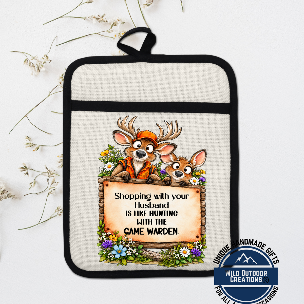 A beige pot holder with a cartoon image of two deer wearing orange clothing and holding a sign that says "Shopping with your husband is like hunting with the game wardens".