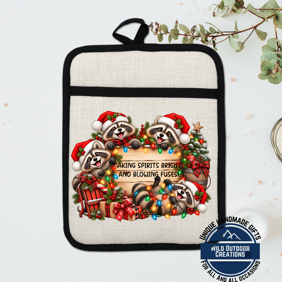 A beige pot holder with a black trim, featuring a festive design of raccoons wearing Santa hats and surrounded by Christmas decorations such as lights, presents, and a wreath.