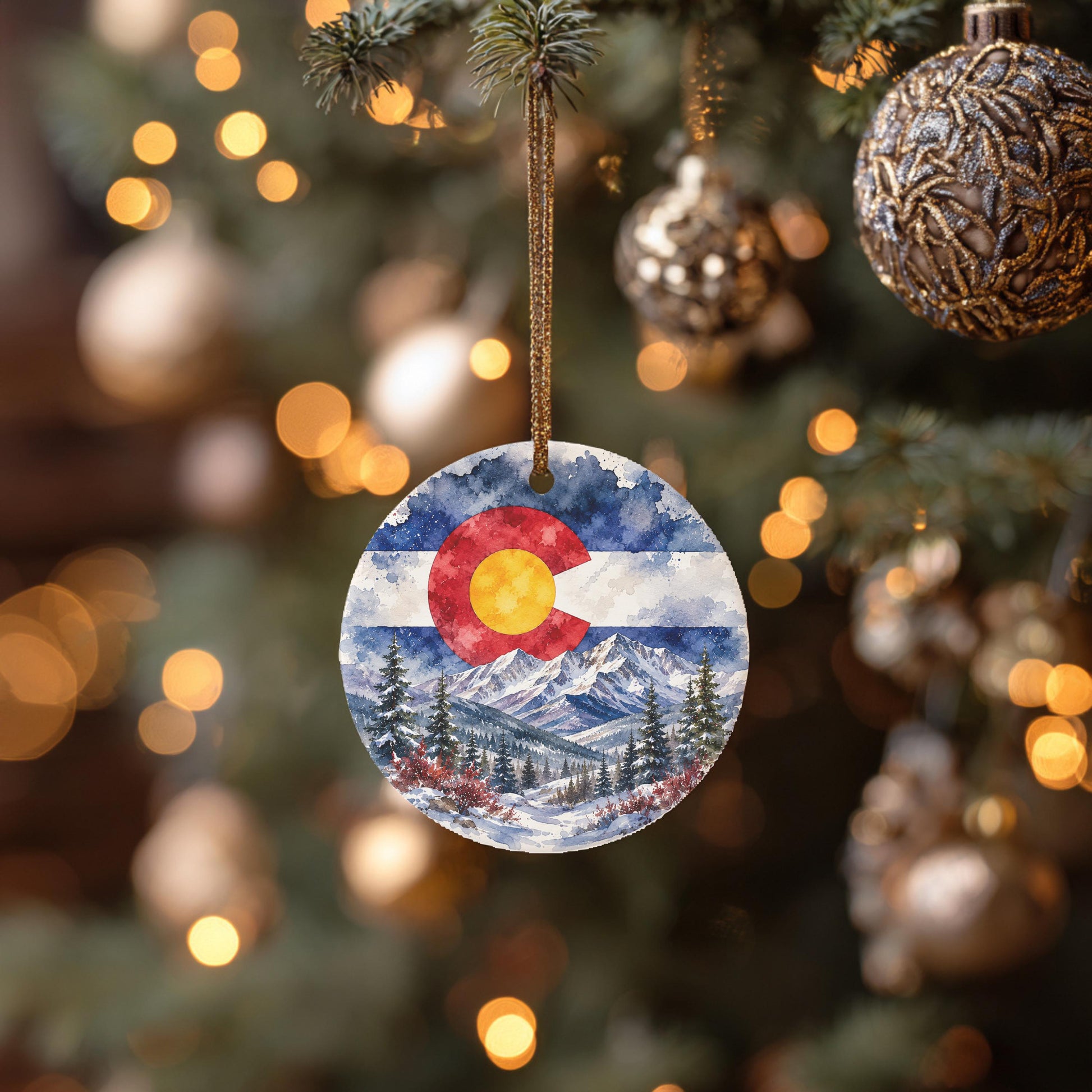A round ornament hanging from a Christmas tree, featuring a snowy mountain landscape with pine trees and a Colorado flag in the center.