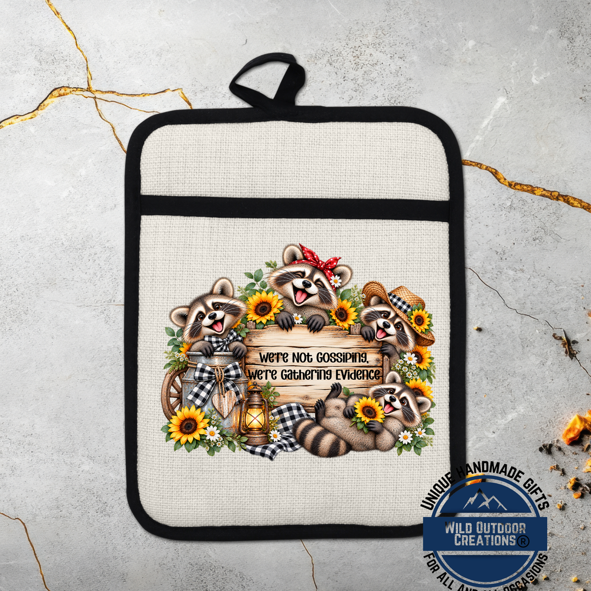 A beige pot holder with a black trim, featuring a design of raccoons surrounded by sunflowers and a sign that says "We're Not Gossipping, We're Gaining Evidence".