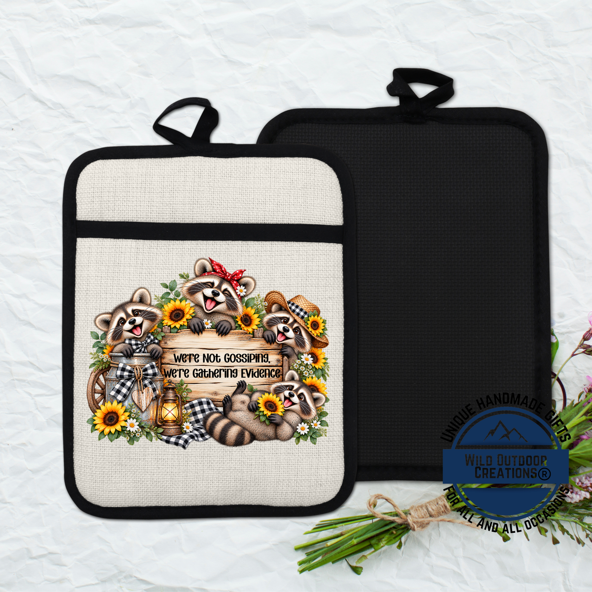 A beige pot holder with a black handle, featuring a design of raccoons surrounded by flowers and a sign that says "We're Not Gossiping We're Gathering Evidence".
