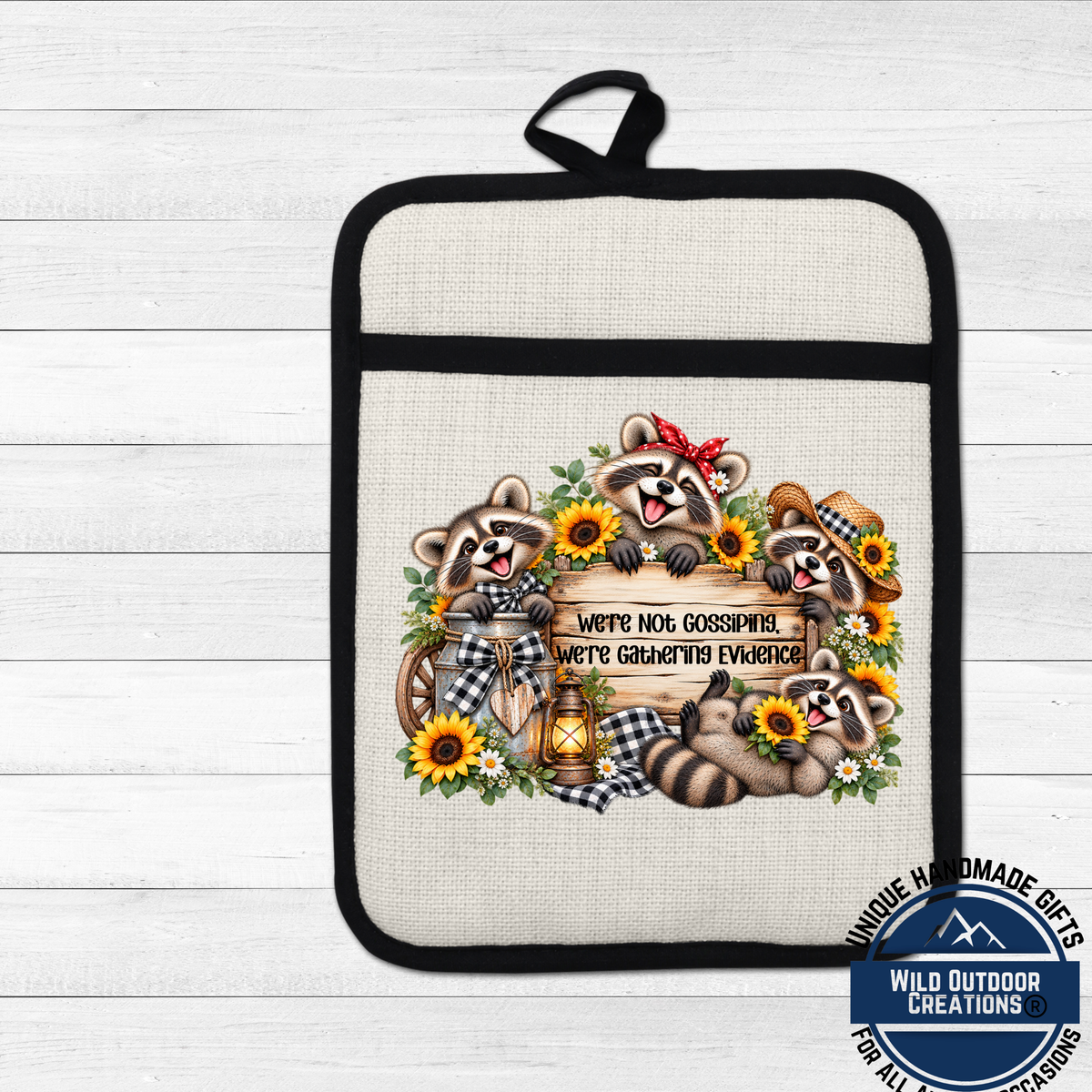 A beige oven mitt with a black handle and a decorative design featuring three raccoons surrounded by sunflowers and a wooden sign with the text "We're Not Gossipping We're Gaining Evidence".