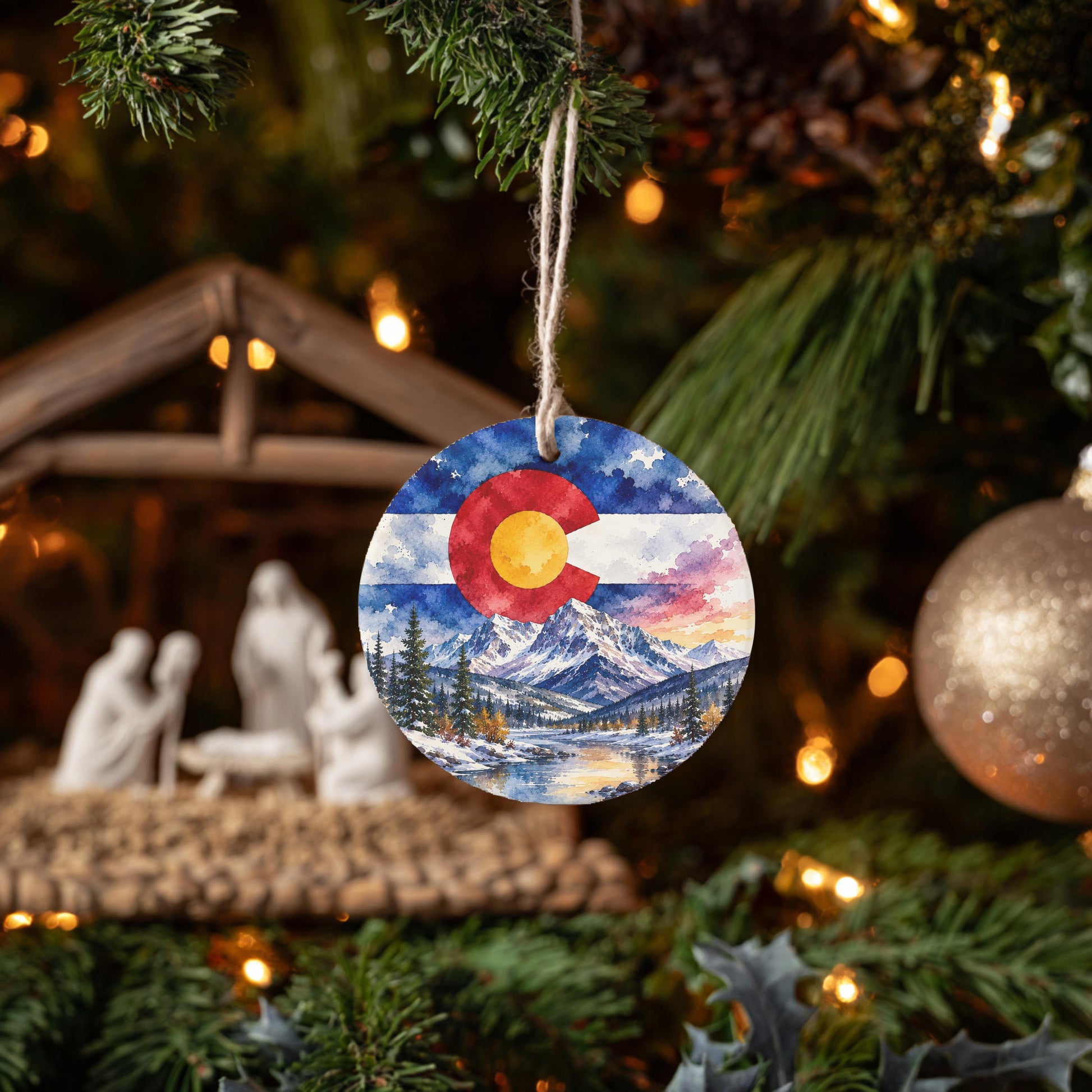 A round ornament with a painting of a mountain landscape hangs from a tree branch, surrounded by other ornaments and lights.