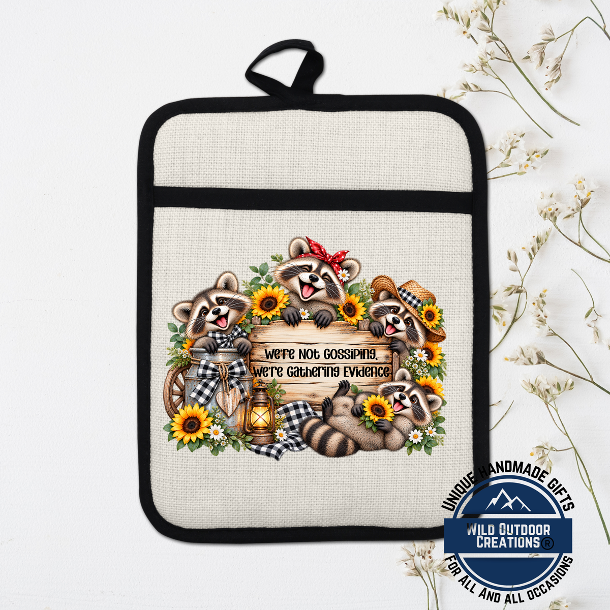 A beige oven mitt with a black handle and a design featuring raccoons surrounded by flowers and a sign that says "We're not gossipping, we're gathering evidence".