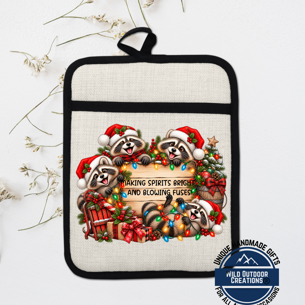 A beige pot holder with a black trim, featuring a festive Christmas wreath design with raccoons wearing Santa hats and surrounded by holiday decorations such as a Christmas tree, presents, and lights.