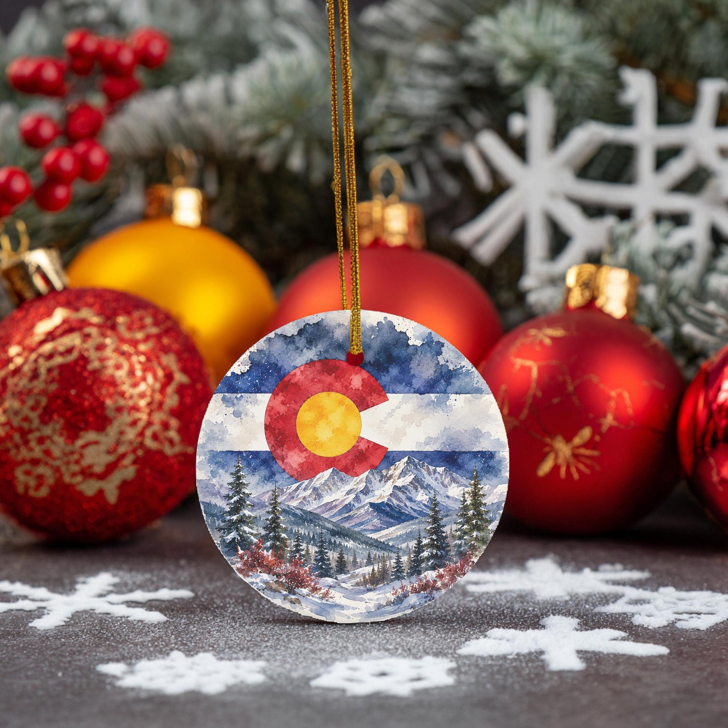 a round ornament with a watercolor painting of a mountain landscape and the Colorado flag, surrounded by various Christmas ornaments and snowflakes.