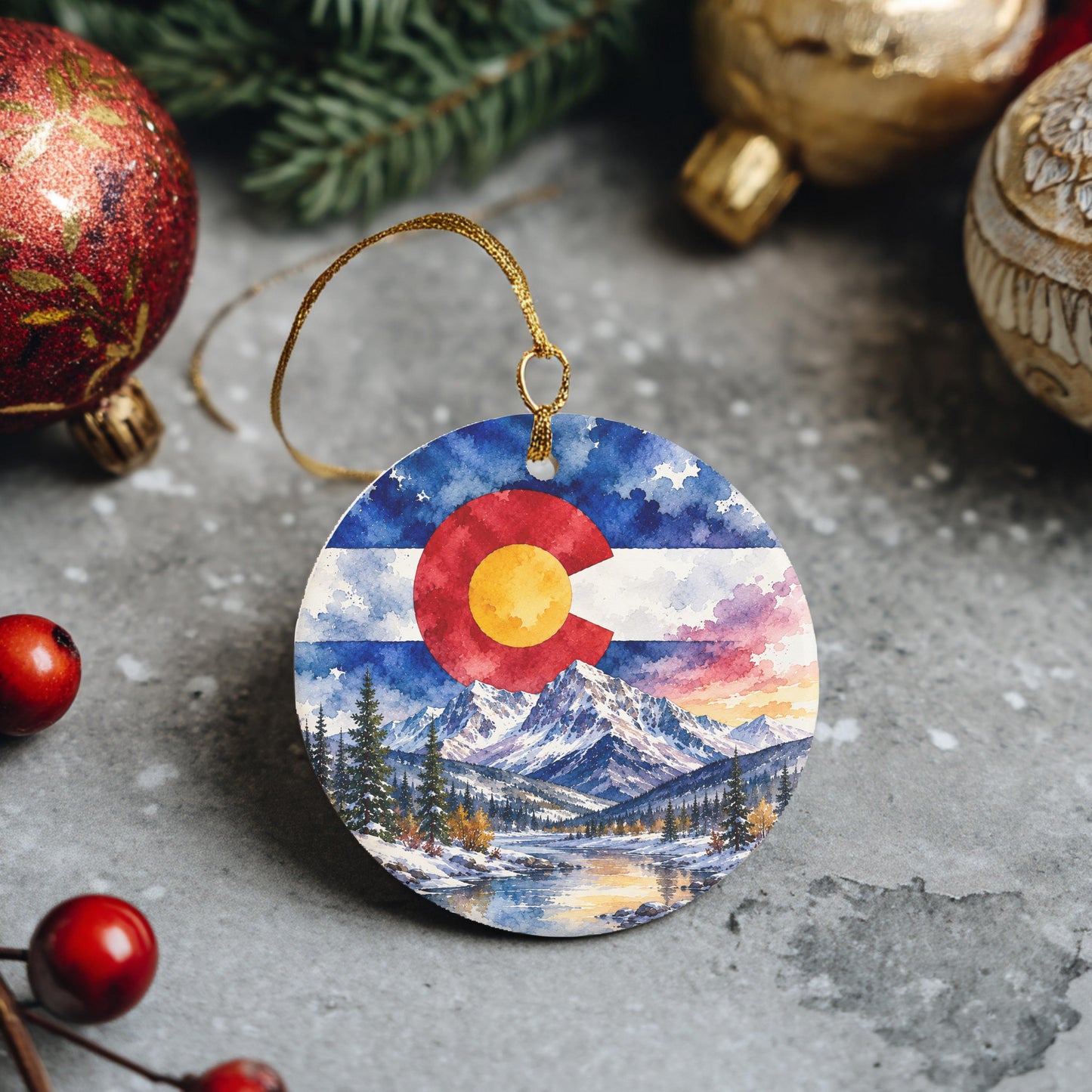A round ornament featuring a painting of a snowy mountain landscape with a Colorado flag in the center, surrounded by Christmas ornaments and berries.