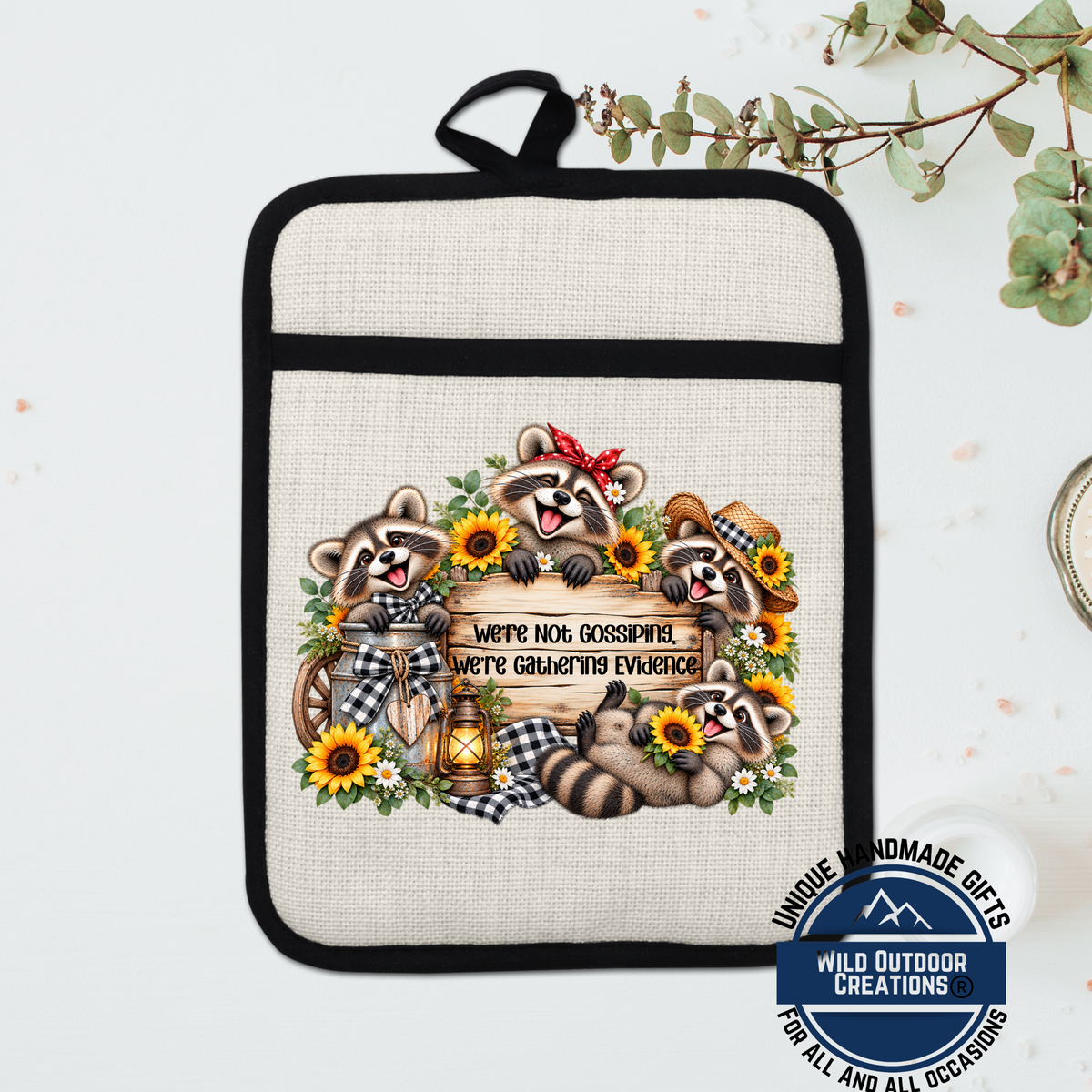 A beige oven mitt with a printed design of raccoons and sunflowers, along with the text "We're not gossipping, we're gathering evidence".