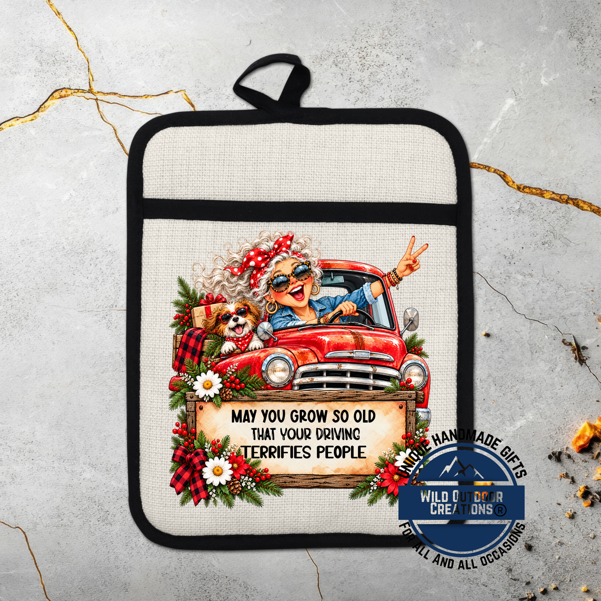 A hand-made pot holder with a cartoon image of an elderly woman driving a red truck, surrounded by flowers and greenery. The text on the pot holder reads "May you grow so old that your driving terrifies people".