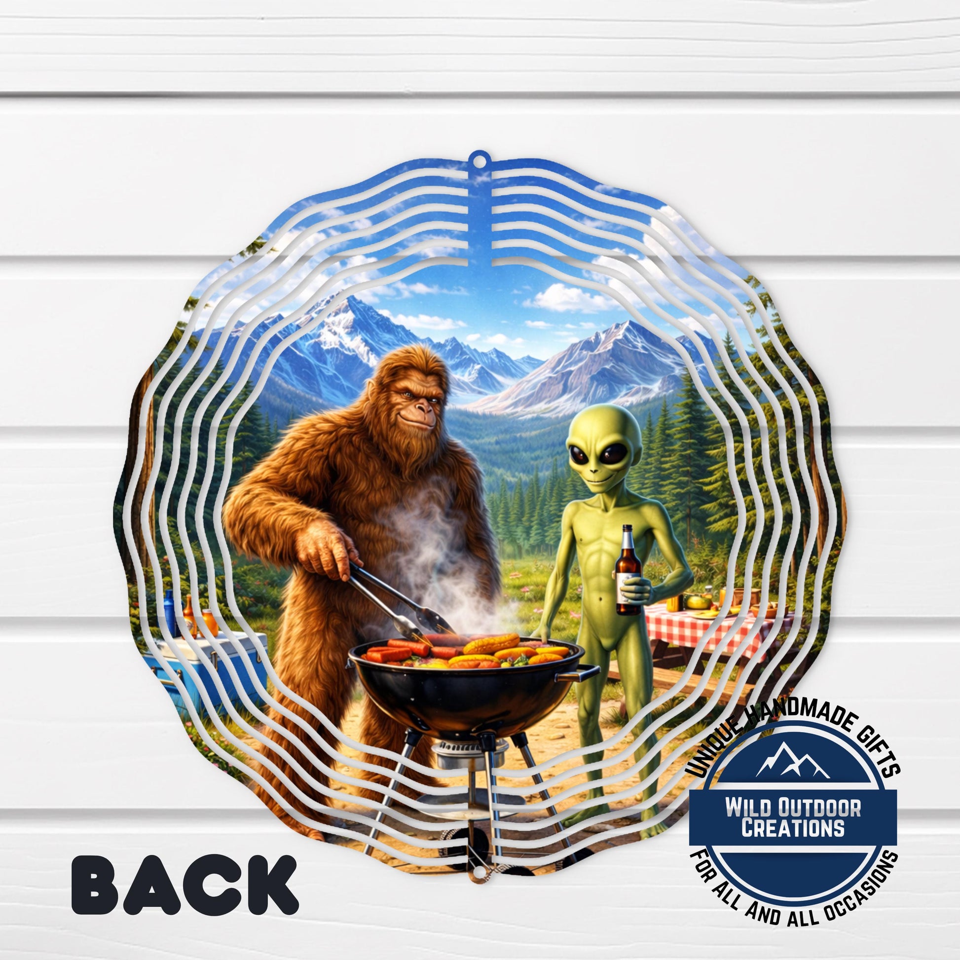 A large metal wind chime with a whimsical scene of a giant brown furry creature cooking food on a grill and an alien holding a bottle, set against a backdrop of mountains and a forest.