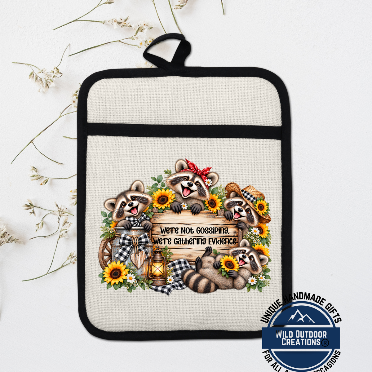 A beige pot holder with a black trim, featuring a whimsical illustration of raccoons surrounded by flowers and a sign that reads "We're Not Gossiping, We're Gaining Evidence".